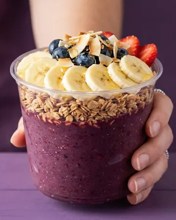 Mão segurando um copo de açaí com banana, mirtilo e morango por cima