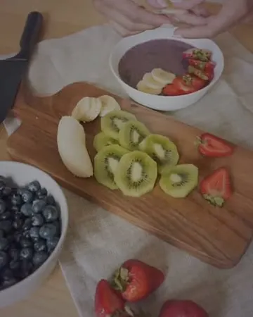 Mãos cortando kiwi, morango e banana na tábua ao lado de uma tigela de açaí